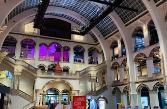 The interior of a large, ornately designed building with arched ceilings and intricate architectural details. The space features gaming machines lined up along the floor, under a high ceiling with a grid pattern. Lighting in purple and white tones enhances the ambiance.