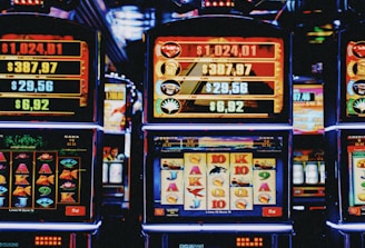 A row of colorful slot machines lit with bright neon lights, showcasing various graphical elements on the screens, indicating different games and jackpots. The atmosphere suggests a lively casino setting, bustling with excitement.