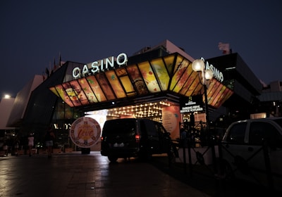 A building with a brightly illuminated entrance is seen at night. The sign above reads 'Casino' in bright letters. The facade features colorful graphics depicting various indulgent themes. Several vehicles are parked near the entrance, and people are visible in the vicinity. Tall lamp posts with spherical lights are present, casting a warm glow.