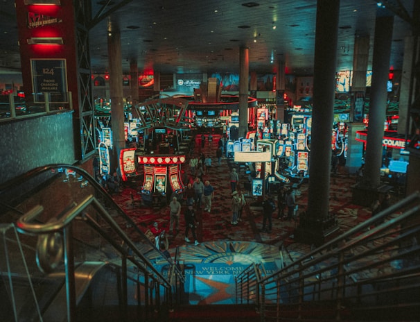 A bustling casino floor filled with groups of slot machines and people walking around. The atmosphere is vibrant with neon lights and illuminated signs. The interior has a ceiling supported by large pillars and there are stairs leading down to the gaming area.